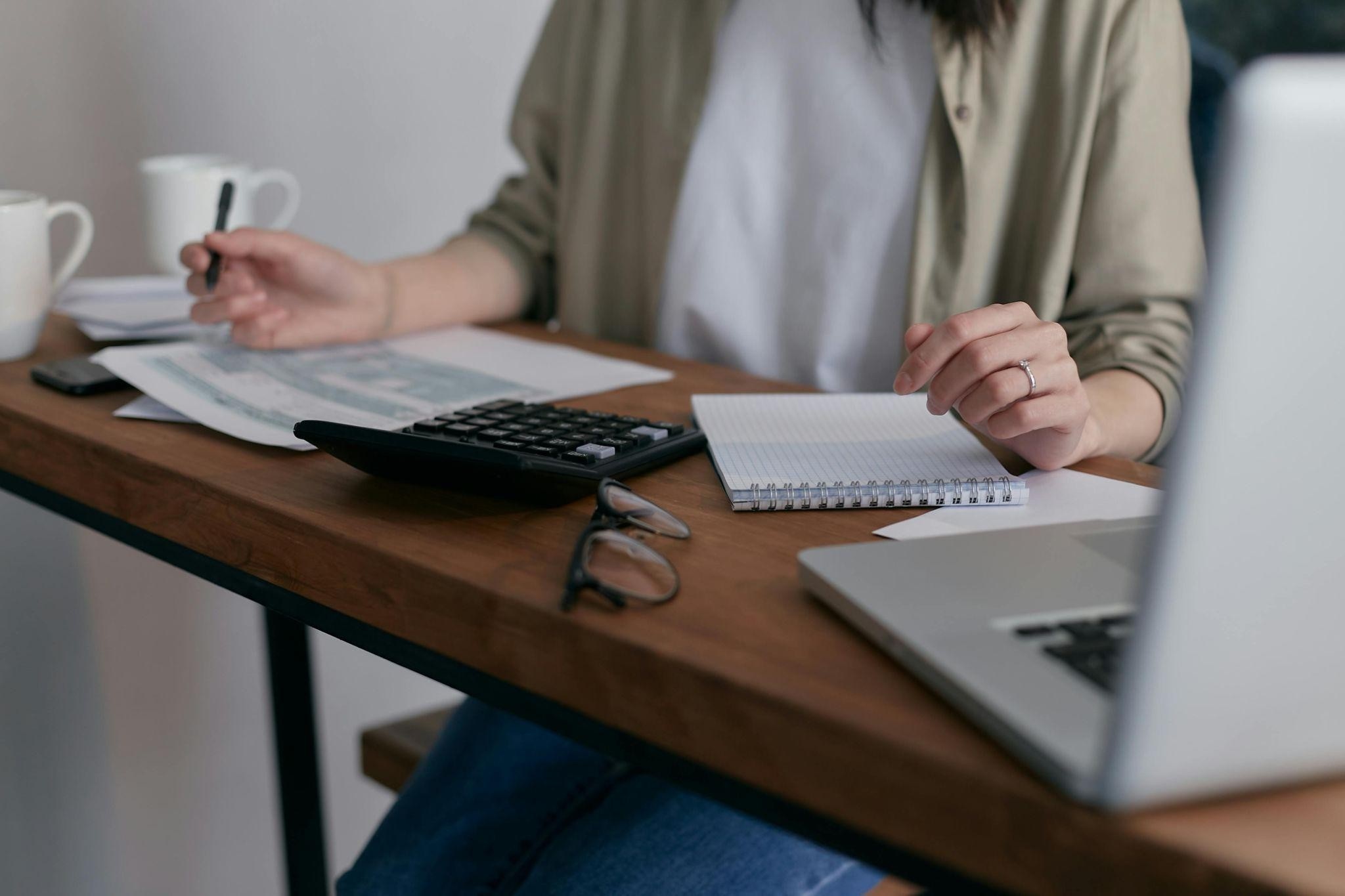 person sitting at a desk with a laptop, calculator, glasses, and a notepad on it